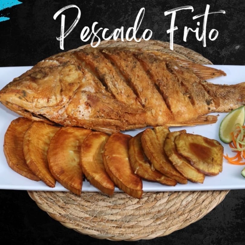 A whole fried fish, Pescado Frito, served with plantain slices and lime wedges on a white plate.